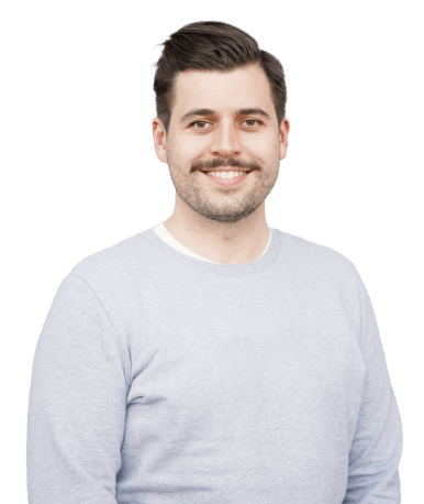 A man with a beard and mustache, wearing a light grey long-sleeve shirt, smiling and posing.