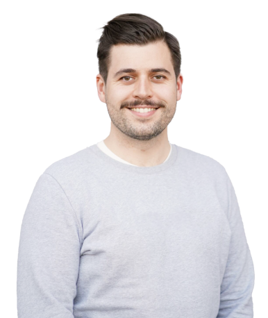 A man with a beard and mustache, wearing a light grey long-sleeve shirt, smiling and posing.