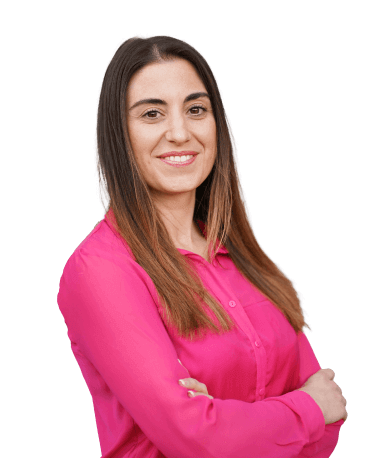 A woman with long hair, smiling and wearing a bright pink blouse, with her arms crossed.